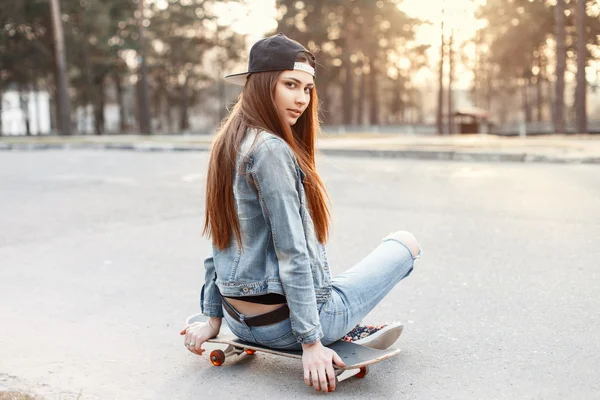 Young beautiful fashionable girl in stylish clothing sitting on a skateboard at sunset