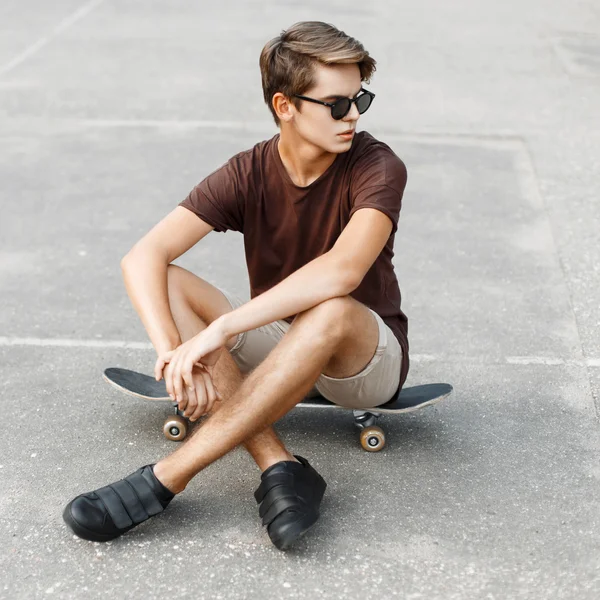 Young stylish man in sunglasses sitting on a skateboard.