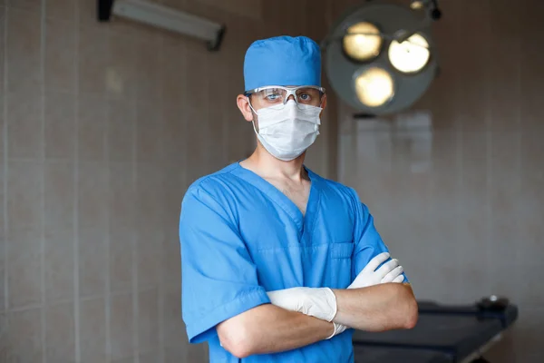 Young professional doctor in a blue uniform, glasses and hat stands in operating room.