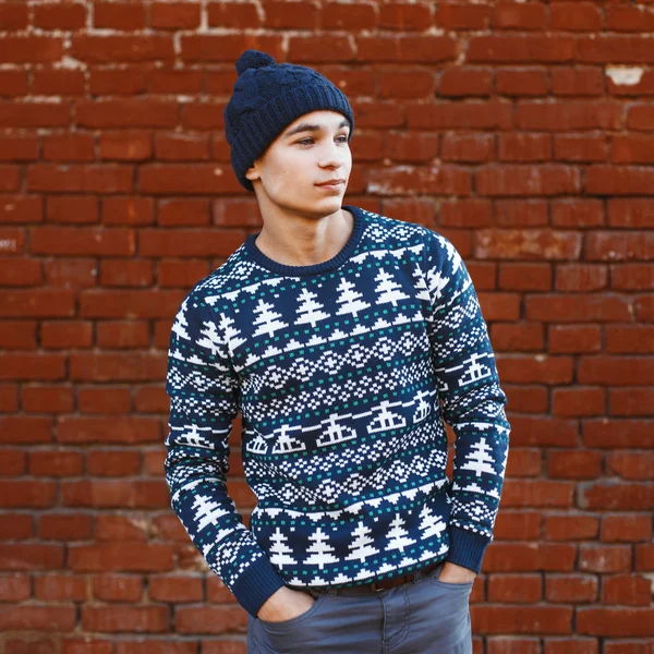 Man in a knitted warm sweater with Christmas patterns and hat standing near red brick wall