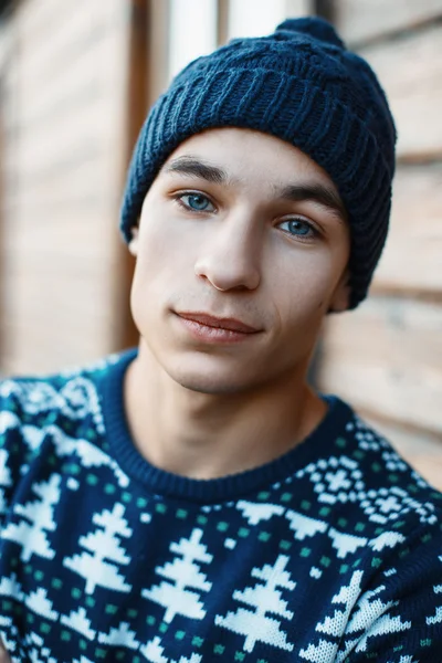 Close-up portrait of a young man in a knitted Christmas sweater and hat on the background of wooden wall.