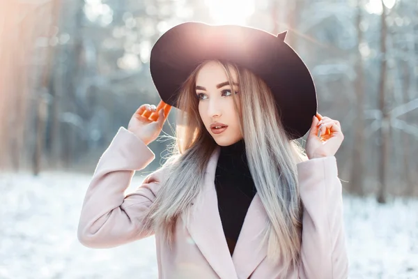 Stylish beautiful woman in elegant hat and coat walking in a winter park at sunset
