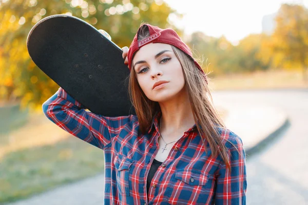Closeup portrait of a young girl in a cap holding a skateboard in the park at sunset.