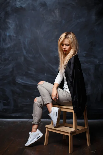 Young beautiful woman with blonde hair in a stylish black shirt, white sneakers and torn jeans sitting on a wooden chair on a dark background.