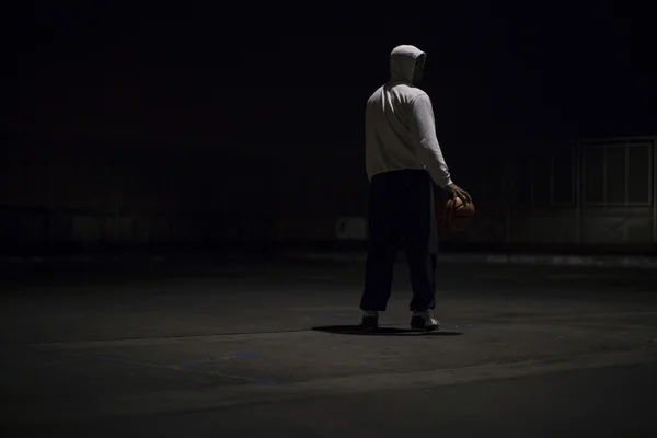 Hooded athlete holds a basketball whilst standing under a street light at night