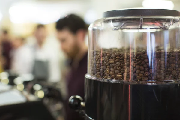 Foreground shallow depth of field of roasted coffee beans in a grinder with blurred activity in the background