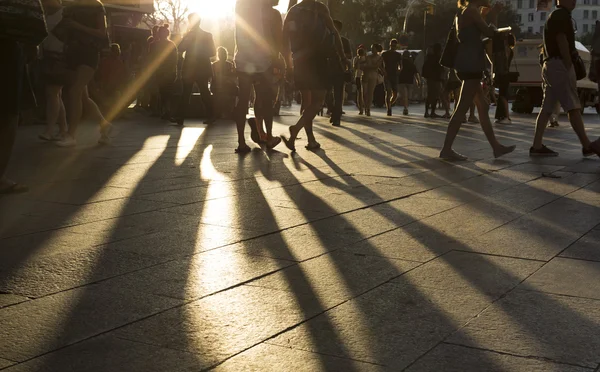 Crowds walking in a busy city district as the sun flares between them in the late afternoon creating long shadows on the ground