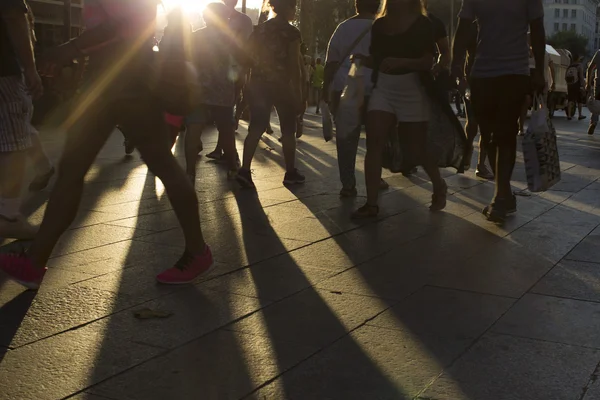 Crowds walking in a busy city district as the sun flares between them in the late afternoon creating shadows in the foreground