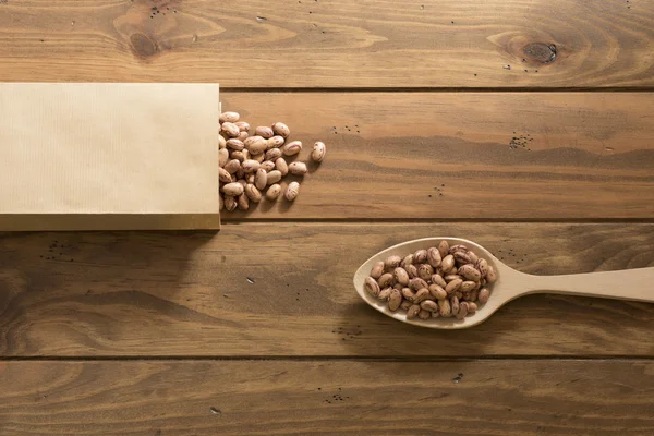A paper bag, some speckled beans and a spoon on a wooden table