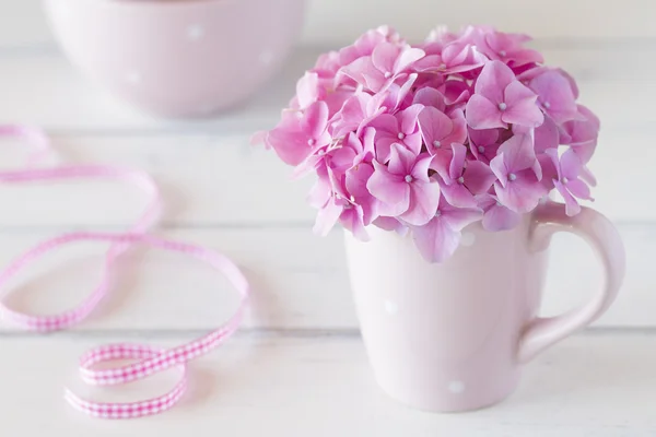 Hydrangea in a cup and a pink checkered ribbon. All on a white wooden table.