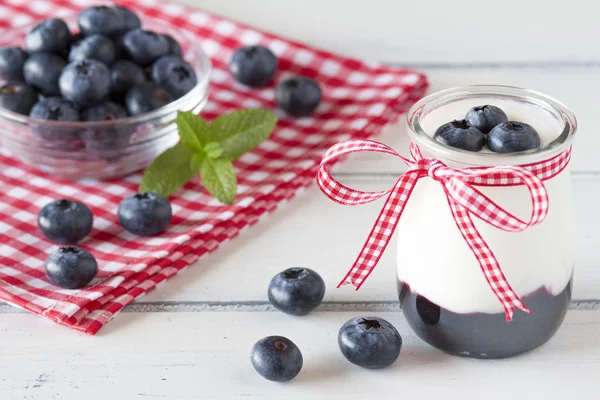 Jar with greek yogurt, blueberry jam and a red ribbon. A bowl off blueberries and peppermint. All in a white wooden table and a checkered red napkin.