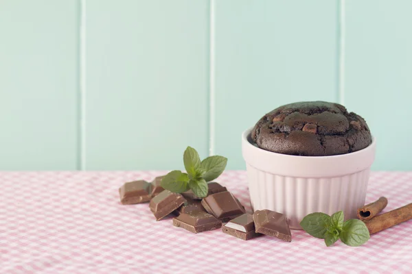 Chocolate muffin in a pink classic whiteware baking bowl, mint, cinnamon and chocolate pieces on a pink checkered tablecloth with a robin egg blue background. Vintage look.