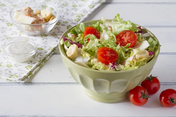 Cesar salad on a green bowl on a white wooden table with ingredients and a napkin
