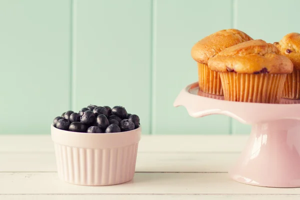Blueberry muffin in a cakestand and several fresh blueberries in a pink classic whiteware baking bowl on a white wooden table with a robin egg blue background. Vintage look.