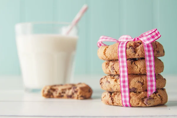 Chocolate chip cookies with a pink ribbon and a glass of milk with a straw on a white wooden table with a robin egg blue background. Vintage look.