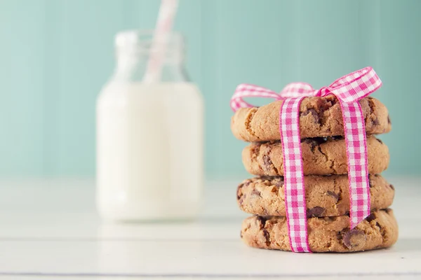 Chocolate chip cookies with a pink ribbon and a school milk bottle with a straw on a white wooden table with a robin egg blue background. Vintage look.
