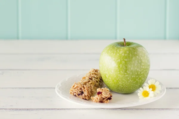 A apple on a plate with honey cereals bar on a white wooden table with a robin egg blue background. Vintage