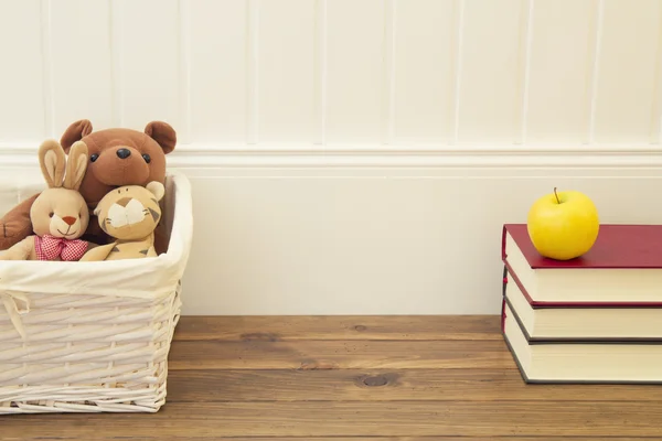 Stuffed animal toys in a basket on the floor. A stack of books and an apple in front of a white wainscot.