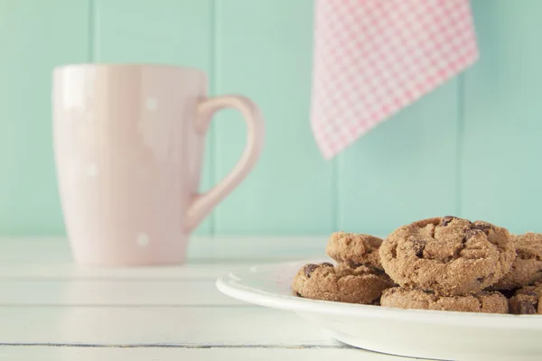 Some chocolate chip cookies on a plate and a pink mug on a white wooden table with a robin egg blue background and a pink checkered napkin. Vintage style.