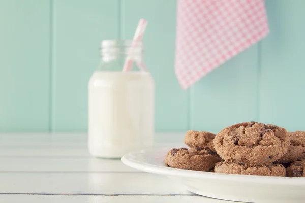 Some chocolate chip cookies on a plate and a school milk bottle with a straw on a white wooden table with a robin egg blue background and a pink checkered napkin. Vintage style.
