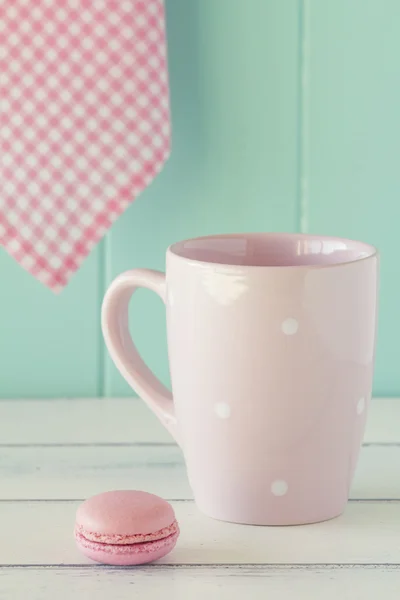 Pink mug with white polka dots, a macaron and a pink checkered napkin on a white wooden table with a robin egg blue background. Vintage Style.