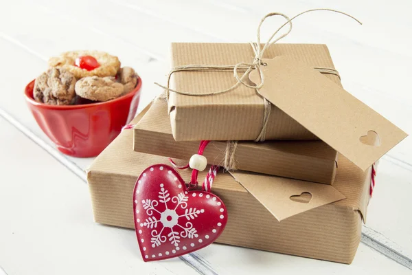 A bowl with cookies, a red heart and some christmas gift boxes wrapped with paper kraft and tied with red & white baker\'s twine on a white wooden table. Vintage Style.
