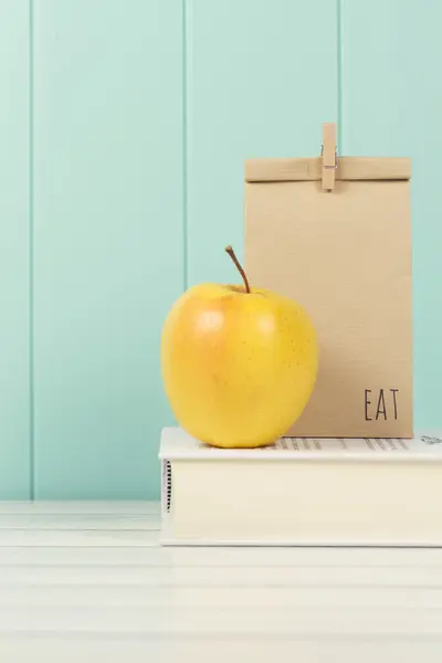 An apple and a paper bag with lunch on a book.