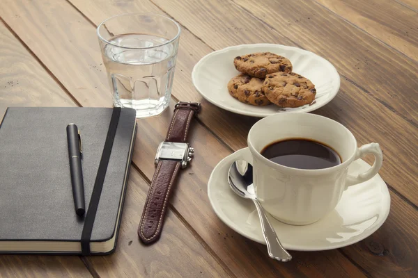 Coffee break at the office. A wooden table with a coffee cup, a notebook, a wristwatch, a water glass and some cookies.