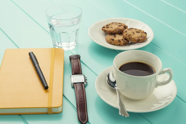 Coffee break at the office. A turquoise wooden table with a coffee cup, a yellow notebook, a wristwatch and a water glass and some cookies.