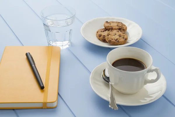 Coffee break at the office. A blue wooden table with a coffee cup, a yellow notebook and a water glass and some cookies.