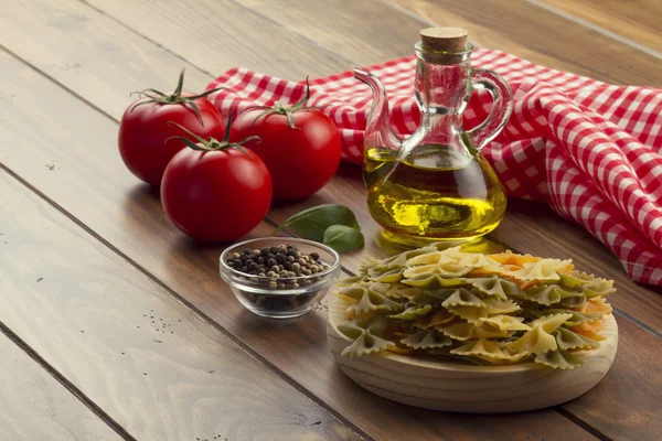 Some ingredients in a wooden table for cooking Italian pasta: tomatoes, farfalle, olive oil, peppermint and pepper. A checkered red napkin.