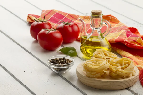 Some ingredients and a napkin in a white wooden table for cooking Italian pasta: tomatoes, nests ribbons pasta, olive oil, peppermint and pepper