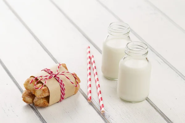 Puff pastry twists sprinkled with sugar with paper kraft and baker\'s twine red white. Two school milk bottles and two straw drink on a white wooden table