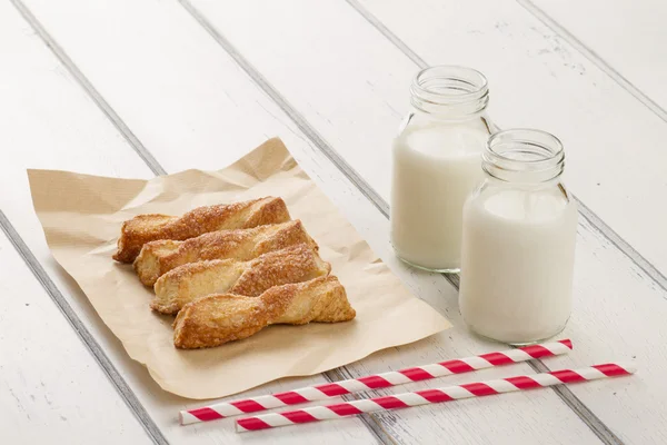 Puff pastry twists sprinkled with sugar on a paper kraft. Two school milk bottles and two straw drink on a white wooden table