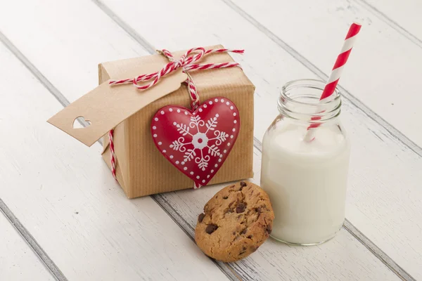 A paper parcel (christmas gift box) with a tag and a red heart, wrapped with paper kraft and tied with red & white baker\'s twine. A chocolate chip cookie and a school milk bottle with a straw on a white wooden table
