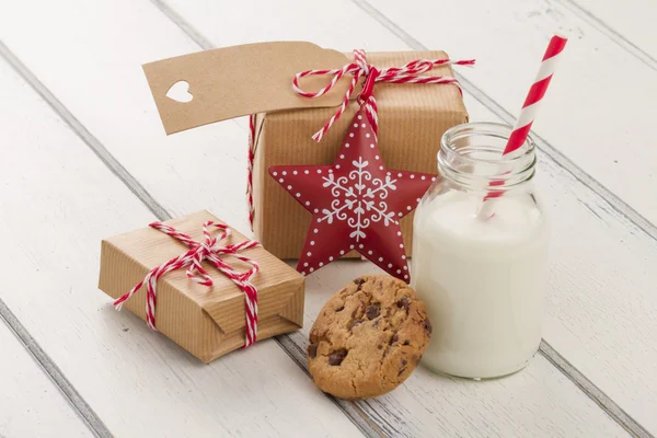 Two paper parcels (christmas gift boxes) with a tag, wrapped with paper kraft and tied with red & white baker\'s twine. A red star, a chocolate chip cookie and a school milk bottle with a straw on a white wooden table