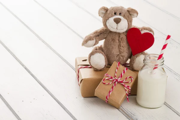 A teddy bear sitting with a red heart. Two paper parcels (christmas gift boxes) wrapped with paper kraft and tied with red & white baker\'s twine. A school milk bottle with a straw on a white wooden table