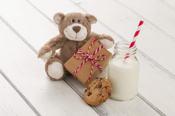 A teddy bear sitting with a paper parcel (christmas gift box) with a tag, wrapped with paper kraft and tied with red & white baker\'s twine. A chocolate chip cookie and a school milk bottle with a straw on a white wooden table