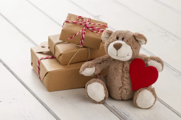 A teddy bear sitting with a red heart. Some paper parcels (christmas gift boxes) wrapped with paper kraft and tied with red & white baker\'s twine on a white wooden table