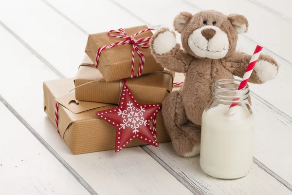 A teddy bear, a red star and a school milk bottle with a straw on a white wooden table. some paper parcels (christmas gift boxes) wrapped with paper kraft and tied with red & white baker's twine.