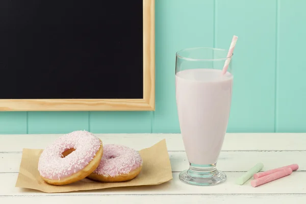 Two delicious strawberry doughnuts on a paper, a chalkboard, some chalks and a strawberry milkshake with a straw on a white wooden table