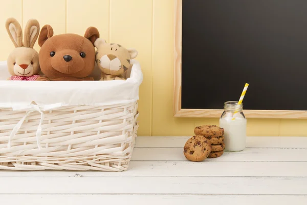 Some stuffed animal toys in a basket, chocolate chip cookies and a school milk bottle with a straw. A chalkboard in the background. Back to school.