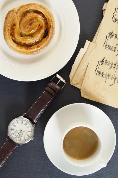 Breakfast: a coffee and a danish puff pastry swirl. A wristwatch and a old music sheet on the table.