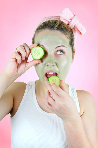 Beauty portrait of a funny woman wearing a pink headband, white top and a green facial mask is holding a slice of cucumber in front of her left eye and is about bite into another one.