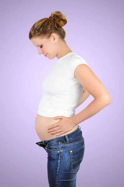 Young woman in early pregnancy wearing a white crop top and jeans is touching her bare baby bump. Purple background.
