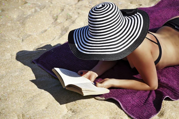 Young elegant woman wearing a black bikini and a large black and white striped beach hat is lying comfortably on the towel on the beach and reading a book.