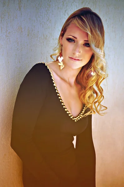 Beautiful young woman with blonde curly hair and smoky eye make-up wearing a black long sleeved dress with golden studs and matching big golden drop earrings.