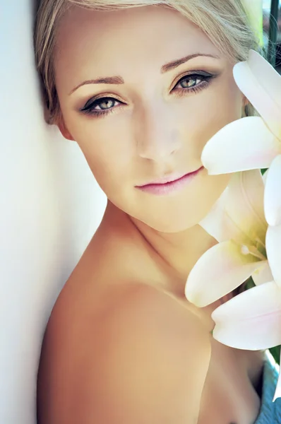Young blonde woman wearing brown eye make-up is leaning against the wall and holding white lilies close to her face.