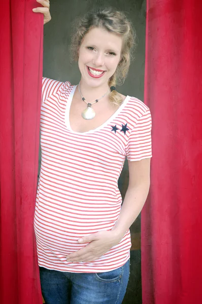 Young smiling pregnant woman with dark hair and eyes wearing red lipstick and a red and white striped T-shirt is standing between dark red curtains and stroking her growing baby bump.