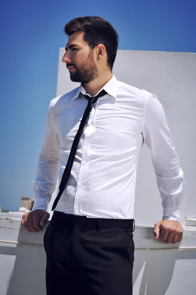 Attractive Spanish looking young man with a beard and dark hair and eyes is wearing a sophisticated white shirt and a black trousers matched with a black tie.